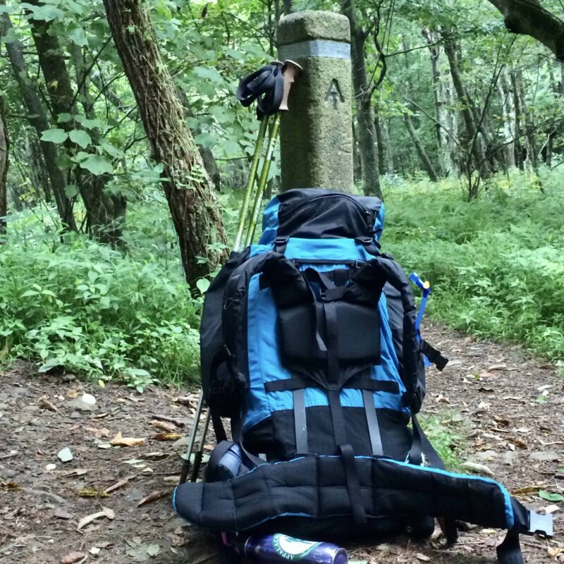 A large blue and black hiking backpack sitting on the ground in the forest against a cement post that has the "AT" symbol representing the Appalachian Trail.